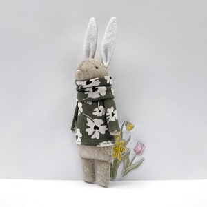 May include: A handmade felt bunny doll wearing a green sweater with white floral print and a matching scarf. The bunny has long white ears and is standing next to a watercolor illustration of daffodils and a tulip.