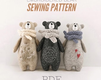 May include: Three plush bears with embroidered details and knit scarves. The left bear is tan with white daisy embroidery, the center is dark gray with white stitch patterns, and the right is light gray with two red hearts. Text reads EMBROIDERED BEAR SEWING PATTERN PDF.