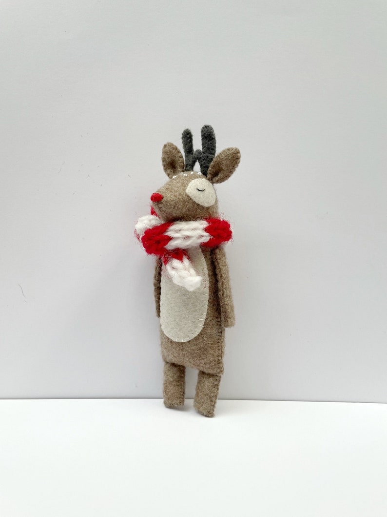 May include: A handmade felt reindeer figurine with a red and white striped scarf. The reindeer has brown fur, a white belly, and small antlers. The figurine is a festive decoration for the holiday season.