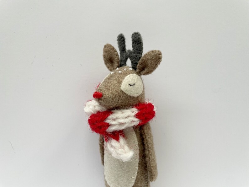 May include: A handmade felt reindeer with a red nose, gray antlers, and a red and white striped scarf. The reindeer has closed eyes and small white details. The overall design is whimsical and festive.