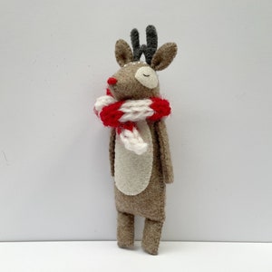 May include: A handmade felt reindeer figurine with a red nose, gray antlers, and a red and white striped scarf. The reindeer has a cream-colored belly and closed eyes, suggesting a whimsical, festive design. This decorative item is likely for holiday decor.