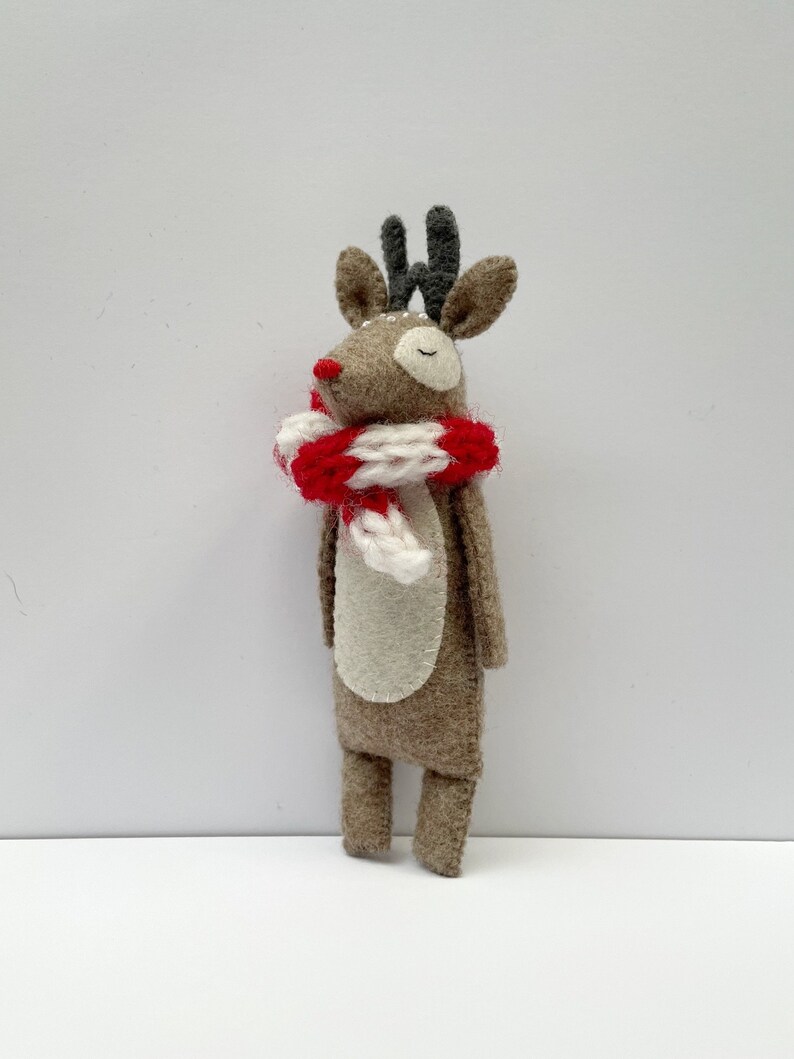 May include: A handmade felt reindeer figurine with a red nose, gray antlers, and a red and white striped scarf. The reindeer has a cream-colored belly and closed eyes, suggesting a whimsical, festive design. This decorative item is likely for holiday decor.