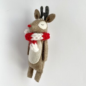 May include: A handmade felt reindeer figurine with a red and white striped scarf. The reindeer has a brown body, gray antlers, and a red nose. The eyes are closed, and the figurine is standing upright.