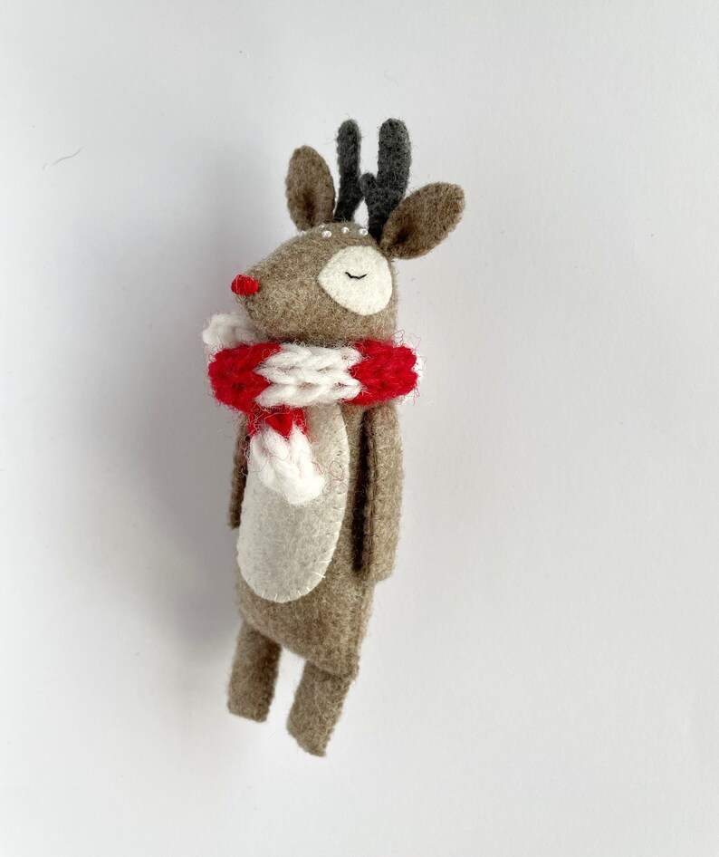 May include: A handmade felt reindeer figurine with a red and white striped scarf. The reindeer has a brown body, gray antlers, and a red nose. The eyes are closed, and the figurine is standing upright.