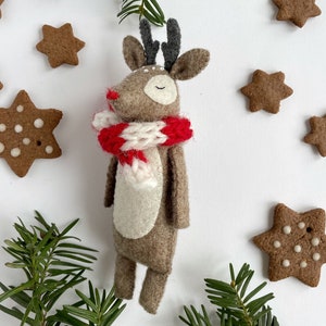 May include: A handmade felt reindeer with a red and white scarf, surrounded by star-shaped gingerbread cookies and evergreen sprigs. The reindeer has closed eyes and a red nose, evoking a festive holiday theme.