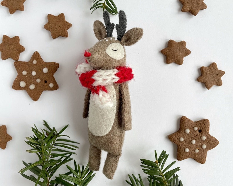 May include: A handmade felt reindeer with a red and white scarf, surrounded by star-shaped gingerbread cookies and evergreen sprigs. The reindeer has closed eyes and a red nose, evoking a festive holiday theme.