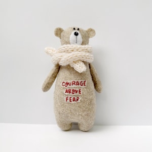 Può includere: Bambola orso in feltro beige, con una sciarpa color crema. L'orso ha dettagli ricamati e le parole "COURAGE ABOVE FEAR" cucite in rosso sul corpo. L'orso è posto su uno sfondo bianco, che mette in risalto il suo design semplice e il suo messaggio.