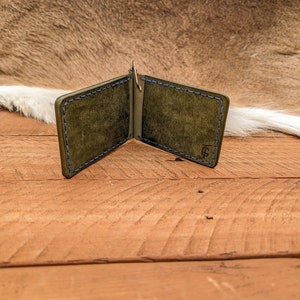 May include: A green leather money clip with blue stitching and a gold metal clip. The money clip is open and lying on a wooden surface.