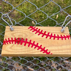 May include: A rectangular wooden sign with a baseball design. The sign is made of light-colored wood and features red stitching details. Two silver carabiner clips are attached to the top for hanging.