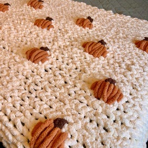 May include: A white, chunky knit blanket with several orange and brown pumpkin appliqués. The blanket is made of a soft, plush material and is perfect for fall decor. The pumpkins are evenly spaced across the blanket.