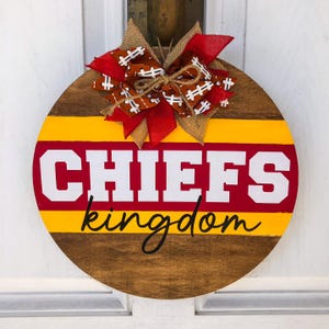 May include: A round wooden door hanger with a red, yellow, and brown striped design. The words "CHIEFS kingdom" are written in white and black lettering. A red and brown bow with football-shaped accents is attached to the top.
