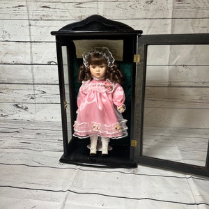 Puede incluir: Una muñeca de porcelana que lleva un vestido rosa con ribete de encaje blanco y un gorro blanco. La muñeca se exhibe en una vitrina de madera negra con frente de vidrio.