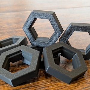 Pode incluir: Um conjunto de seis anéis hexagonais de plástico preto, empilhados e sobrepostos. Os anéis têm um acabamento liso e fosco e uma borda ligeiramente elevada.