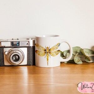 May include: White ceramic mug with a watercolor illustration of a dragonfly with yellow and brown wings. The mug is sitting on a wooden table with a camera and green leaves.