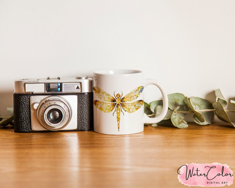 May include: White ceramic mug with a watercolor illustration of a dragonfly with yellow and brown wings. The mug is sitting on a wooden table with a camera and green leaves.