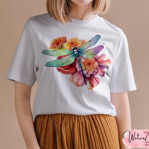 May include: White t-shirt with a watercolor design of a dragonfly and flowers. The dragonfly is blue, green, and purple, and the flowers are pink, orange, and yellow.