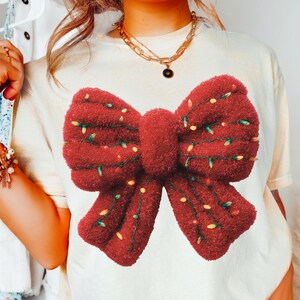 May include: Cream-coloured t-shirt featuring a large, textured red bow graphic adorned with small, yellow and green lights. The bow is the focal point, suggesting a festive or holiday theme. The shirt is worn with blue jeans.