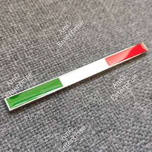 Pode incluir: Um emblema metálico retangular com as cores verde, branco e vermelho, assemelhando-se a uma bandeira. O emblema tem uma superfície brilhante e reflexiva e está colocado sobre um fundo cinzento texturizado. O texto "AmG Enterprise" está impresso no fundo.