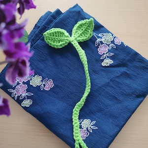 Könnte beinhalten: Ein handgefertigter grüner Häkel-Lesezeichen in Form eines Sprosses mit zwei Blättern oben. Das Lesezeichen liegt auf einem gefalteten marineblauen Stoff mit floraler Stickerei. Im Hintergrund sind lila Blumen zu sehen.