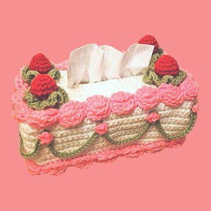 May include: A crocheted tissue box cover in the shape of a cake. The cover is white with pink frosting and decorative pink roses. Red crocheted strawberries with green leaves adorn the top. White tissues peek out from the top.