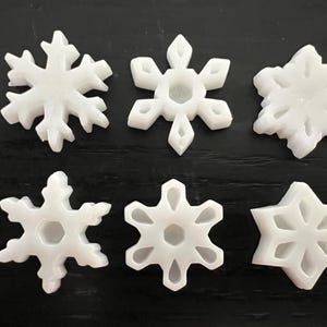 May include: Six white snowflake ornaments in various designs are arranged on a dark surface. Each snowflake has a unique shape, with intricate details and cutouts. The ornaments are made of a solid, opaque material.