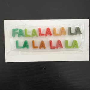 May include: A package of colorful gummy candies spelling out "FALALALA LA LA LA" in various colors. The candies are in the shape of letters and are packaged in a clear plastic wrapper on a white card.