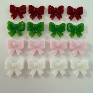 May include: An assortment of sixteen bow-shaped embellishments in four colors: red, green, pink, and white. Each color has four bows arranged in a grid pattern. The bows have a glossy, smooth finish.