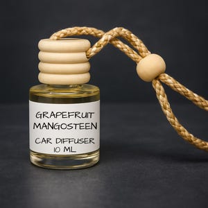 May include: A clear glass car air freshener bottle with a wooden cap and a braided rope. The label reads "GRAPEFRUIT MANGOSTEEN CAR DIFFUSER 10 ML". The liquid inside is yellow. The background is dark gray.