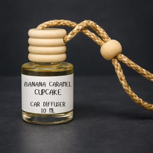 May include: A clear glass car air freshener bottle with a wooden cap and a braided rope. The label reads "Banana Caramel Cupcake Car Diffuser 10 ML". The liquid inside is light yellow. The background is dark gray.