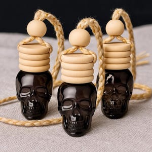 May include: Three black skull-shaped car air fresheners with wooden lids and hanging cords. The lids are light brown, and the cords are a beige color. The air fresheners are displayed on a light gray surface.