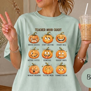 May include: A light green t-shirt featuring a graphic titled TEACHER MOOD CHART with nine pumpkins displaying different facial expressions and labels including INDOOR RECESS, STAFF MEETING, FRIDAY BELL, BEFORE COFFEE, DISMISSAL, PARENT EMAIL, MONDAY MORNING, TESTING WEEK, and SUMMER BREAK.
