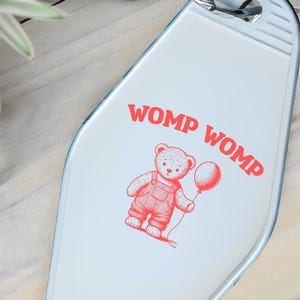 Puede incluir: Un llavero azul claro estilo motel con una ilustración roja de un oso de peluche sosteniendo un globo. El texto "WOMP WOMP" está impreso en rojo encima del oso. El llavero tiene un anillo de metal plateado.