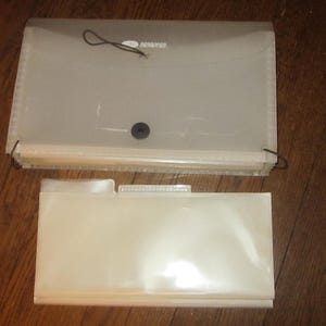 May include: Two clear plastic expanding file folders on a wooden surface. The top folder is closed with a black elastic cord and button closure. The bottom folder is open, with a tab for labeling.
