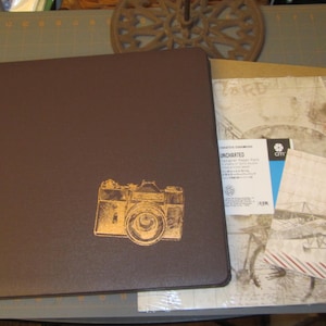 May include: A brown leather photo album with a gold embossed camera design. The album is closed and has a textured surface. The album is sitting on a table with a piece of paper with a vintage design.