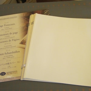 May include: An open scrapbook with cream-colored pages sits next to a package of page protectors. The package is labeled "Page Protectors" in multiple languages, including English, French, and Spanish. The page protectors are for 12 x 12 inch pages.