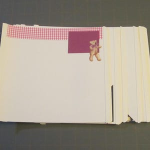Könnte beinhalten: Ein Stapel weißer Papierblätter, die mit Heftklammern zusammengebunden sind. Ein rosa-weiß kariertes Klebeband und ein weinrotes Quadrat sind oben angebracht. Ein kleiner Teddybär-Aufkleber befindet sich auf dem weinroten Quadrat.