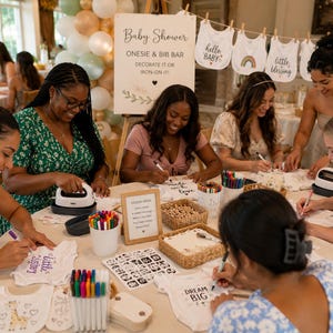 Puede incluir: Una estación de decoración de bodies y baberos para baby shower con prendas blancas, marcadores de tela coloridos, prensas térmicas y un letrero que dice Baby Shower Onesie & Bib Bar Decorate It Or Iron-On It con tarjetas de ideas.