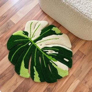 Puede incluir: Una alfombra con forma de hoja de monstera verde y blanca sobre un suelo de madera. La alfombra está hecha de hilo esponjoso y tiene un efecto 3D.