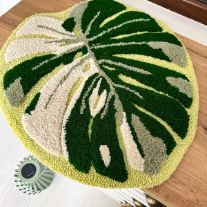 May include: A round, green and cream-colored rug with a monstera leaf design. The rug is made of yarn and has a fluffy texture.
