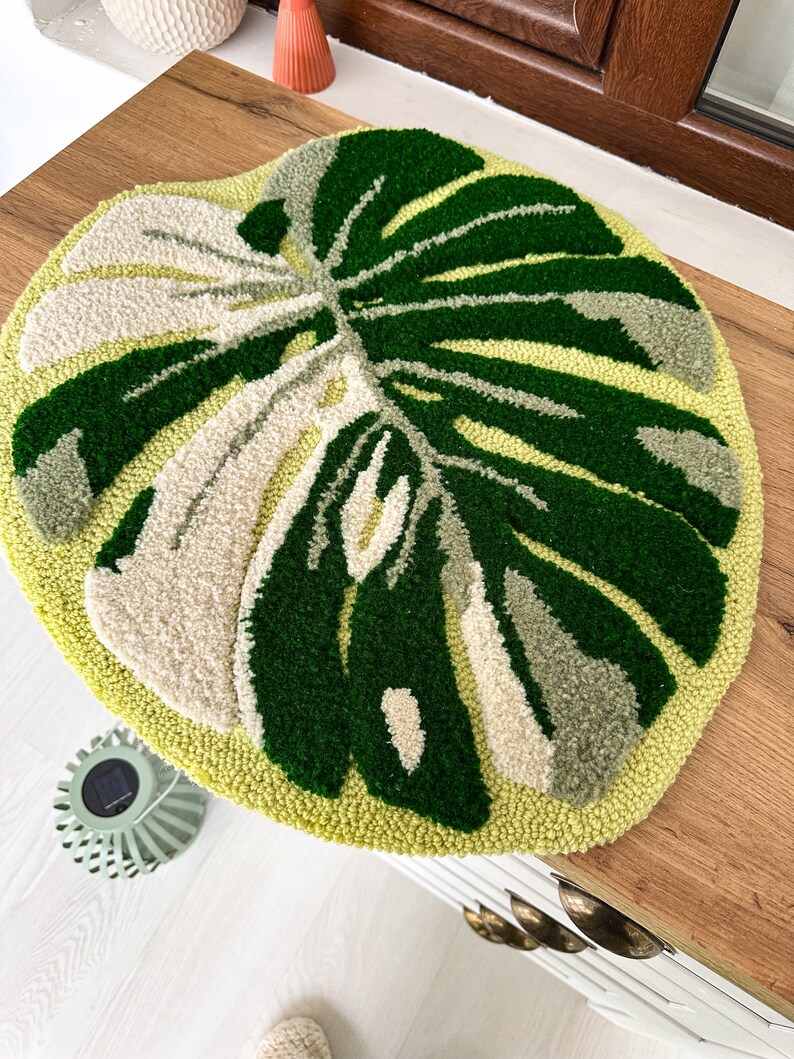May include: A round, green and cream-colored rug with a monstera leaf design. The rug is made of yarn and has a fluffy texture.