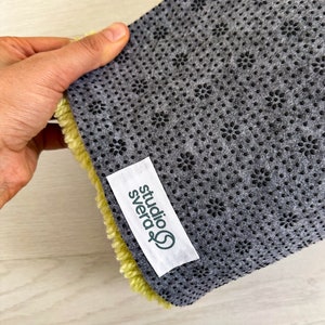 May include: Close-up of the back of a yellow rug with a gray non-slip backing. The backing has a pattern of small black flowers. A white label with green text reads "studio svera".