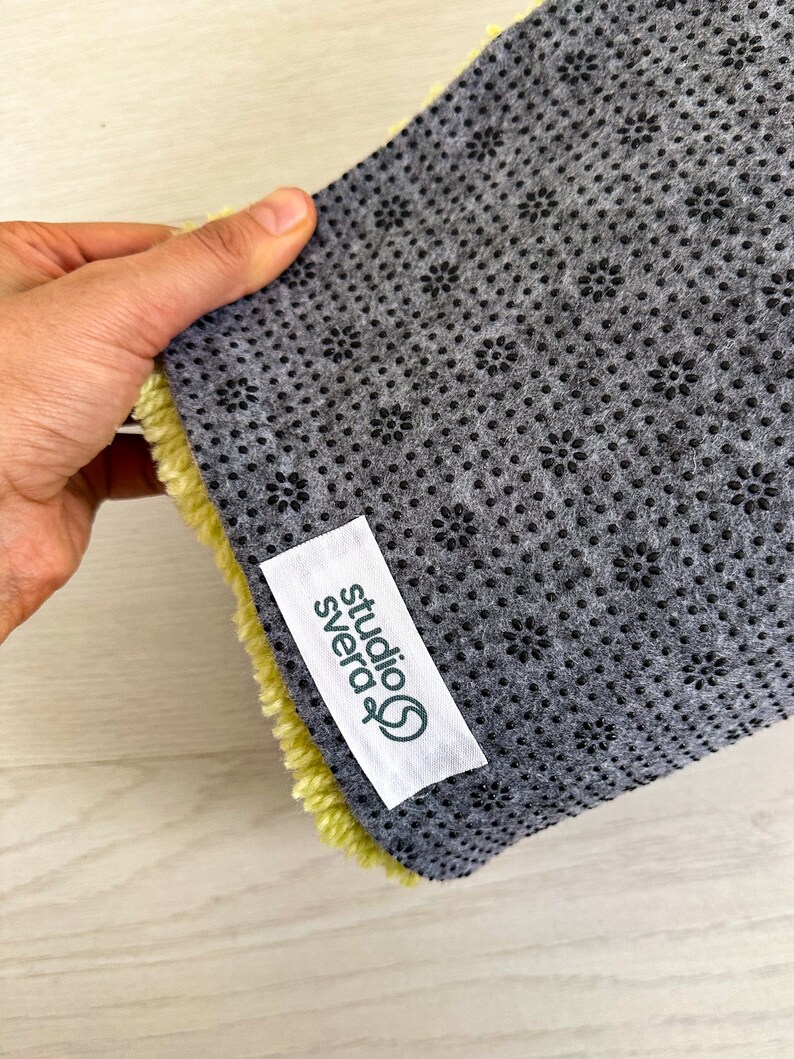 May include: Close-up of the back of a yellow rug with a gray non-slip backing. The backing has a pattern of small black flowers. A white label with green text reads "studio svera".
