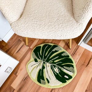 May include: A round, green and white rug with a large leaf design. The rug is on a wood floor in front of a white chair with gold legs.