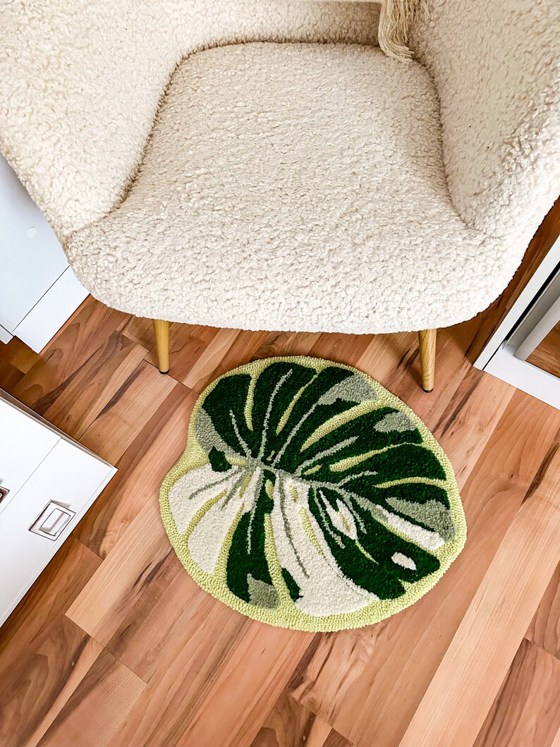 May include: A round, green and white rug with a large leaf design. The rug is on a wood floor in front of a white chair with gold legs.