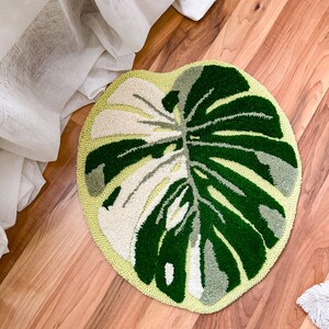 May include: A green and white round rug with a monstera leaf design. The rug is on a wooden floor.