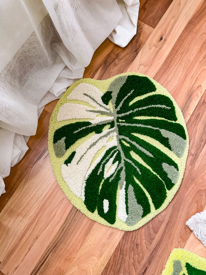 May include: A green and white round rug with a monstera leaf design. The rug is on a wooden floor.