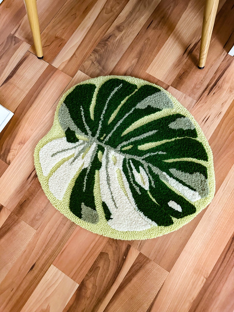 May include: A round, green and white rug with a monstera leaf design. The rug is on a wood floor.