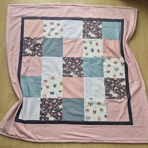 Könnte beinhalten: Eine Patchwork-Decke mit einem rosa gepunkteten Rand und einer dunkelgrauen Umrandung. Die Mitte der Decke zeigt Quadrate aus verschiedenen Stoffen, darunter Rosa, Weiß, Grau und Blau, mit Einhorn- und Tierdrucken.