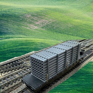 May include: A model train car carrying a load of gray metal pipes. The car is on a set of train tracks, and the background is a green grassy field.