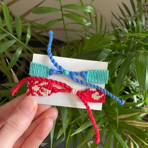 Peut inclure: Trois bracelets d'amiti&eacute; faits &agrave; la main dans des tons de bleu, turquoise et rouge. Les bracelets sont pr&eacute;sent&eacute;s sur une carte blanche sur fond de feuillage vert. Les bracelets sont faits de fils tiss&eacute;s et pr&eacute;sentent des motifs complexes.